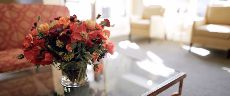 A close-up of a glass vase filled with a colorful bouquet of red, orange, and yellow flowers placed on a glass coffee table in a softly lit living room with beige armchairs and a patterned sofa in the background.