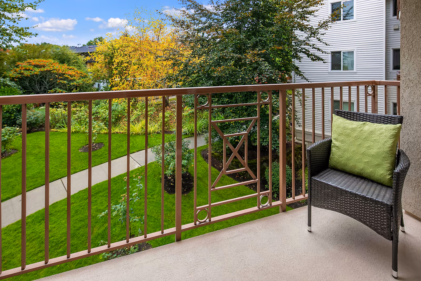 View from a balcony with a brown metal railing overlooking a green garden with a paved walkway, trees with green and yellow leaves, and a wicker chair with a green cushion on the balcony.