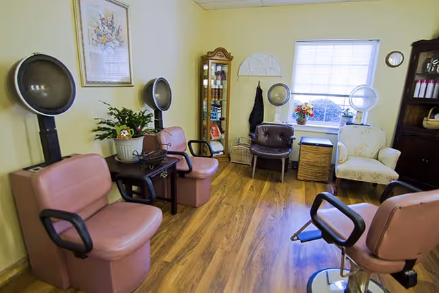 Small hair salon room with pink styling chairs, hooded dryers, wooden floor, and a windowed seating area.