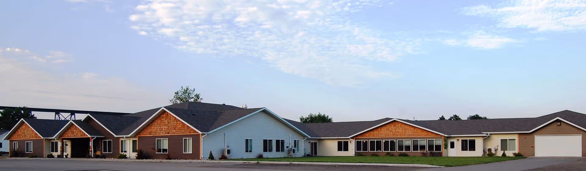 Wide exterior view of a single-story senior living facility building with multiple sections featuring brown and white siding, large windows, and a garage, under a partly cloudy sky.