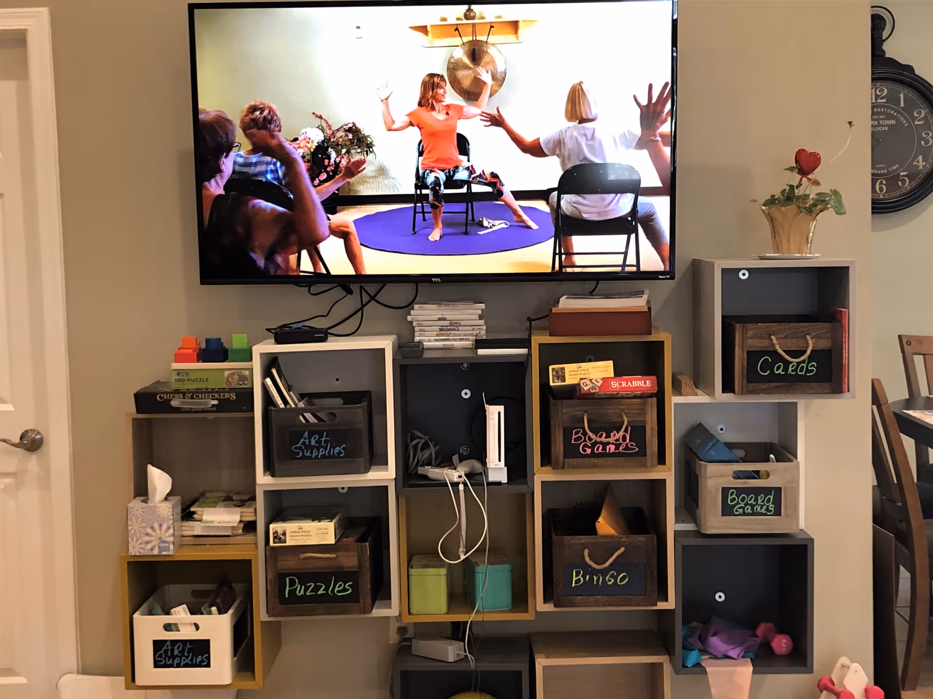 A wall-mounted television screen showing a group of people participating in a seated exercise or yoga session. Below the TV is a shelving unit with labeled wooden and plastic bins containing art supplies, puzzles, board games, cards, and bingo materials. A clock and a small plant are visible on the wall to the right.
