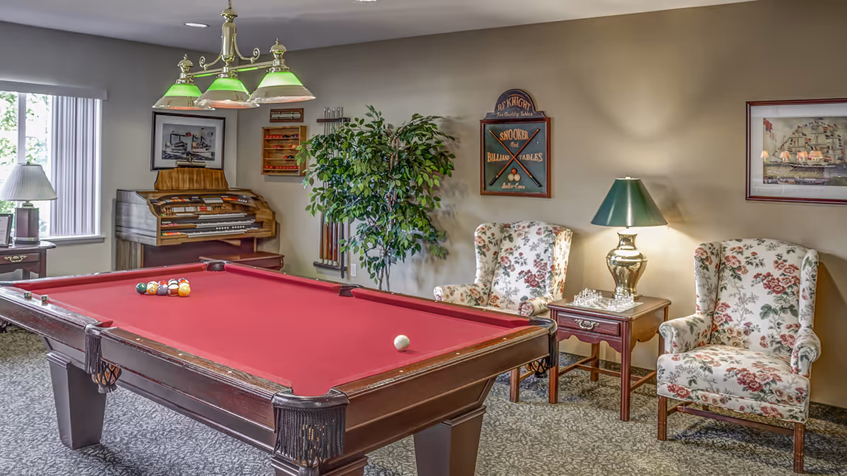 A recreational room with a red felt pool table in the center, two floral upholstered armchairs, a side table with a lamp and a chess set, a large potted plant, a wall-mounted rack holding pool cues, and a vintage organ near a window with vertical blinds.