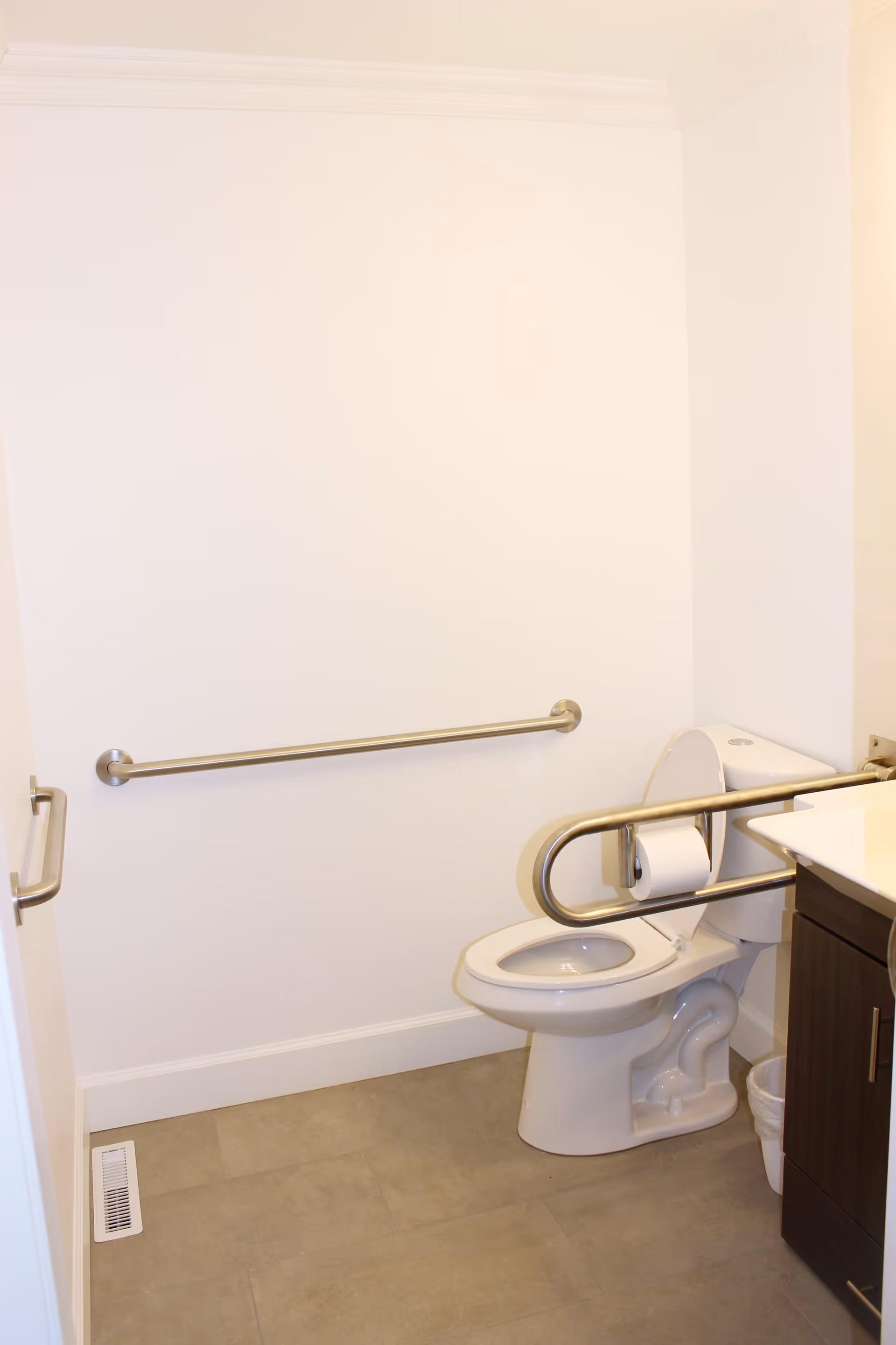 A clean, accessible bathroom with a white toilet equipped with safety grab bars on both sides, a toilet paper holder attached to one grab bar, a white wall with a horizontal grab bar, a dark wood vanity with a white sink, and a small trash bin on the floor.