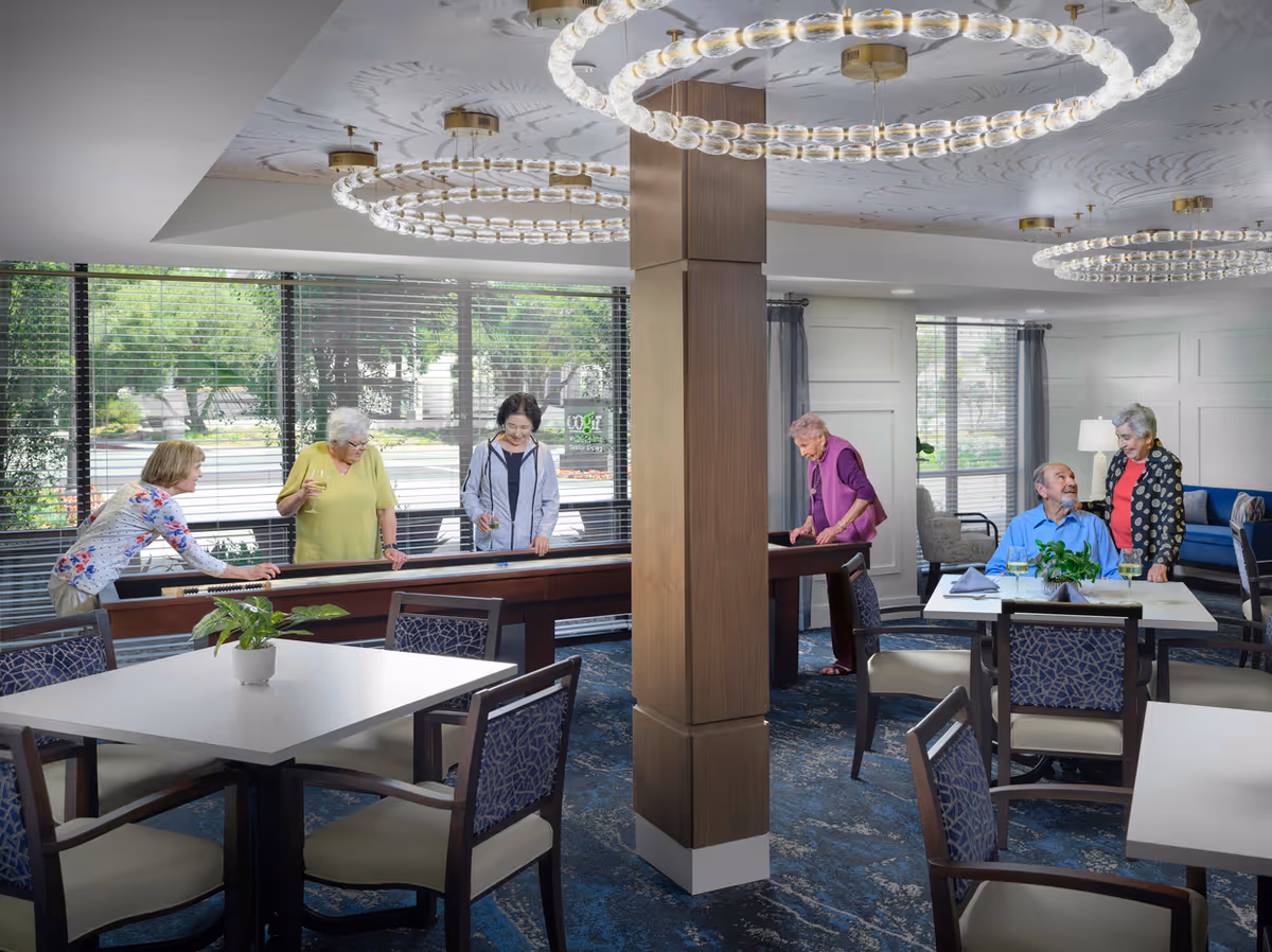 A group of elderly people socializing and playing shuffleboard in a well-lit common area with large windows, modern circular ceiling lights, and tables with chairs arranged around the room. Some are standing and playing the game, while others are seated and conversing.