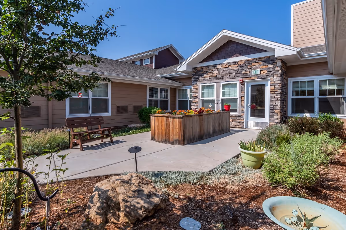 Outdoor courtyard area at Willowbrook Place featuring a wooden bench, a raised wooden planter box with flowers, various green plants and shrubs, a stone and siding building with multiple windows and a glass door under a clear blue sky.