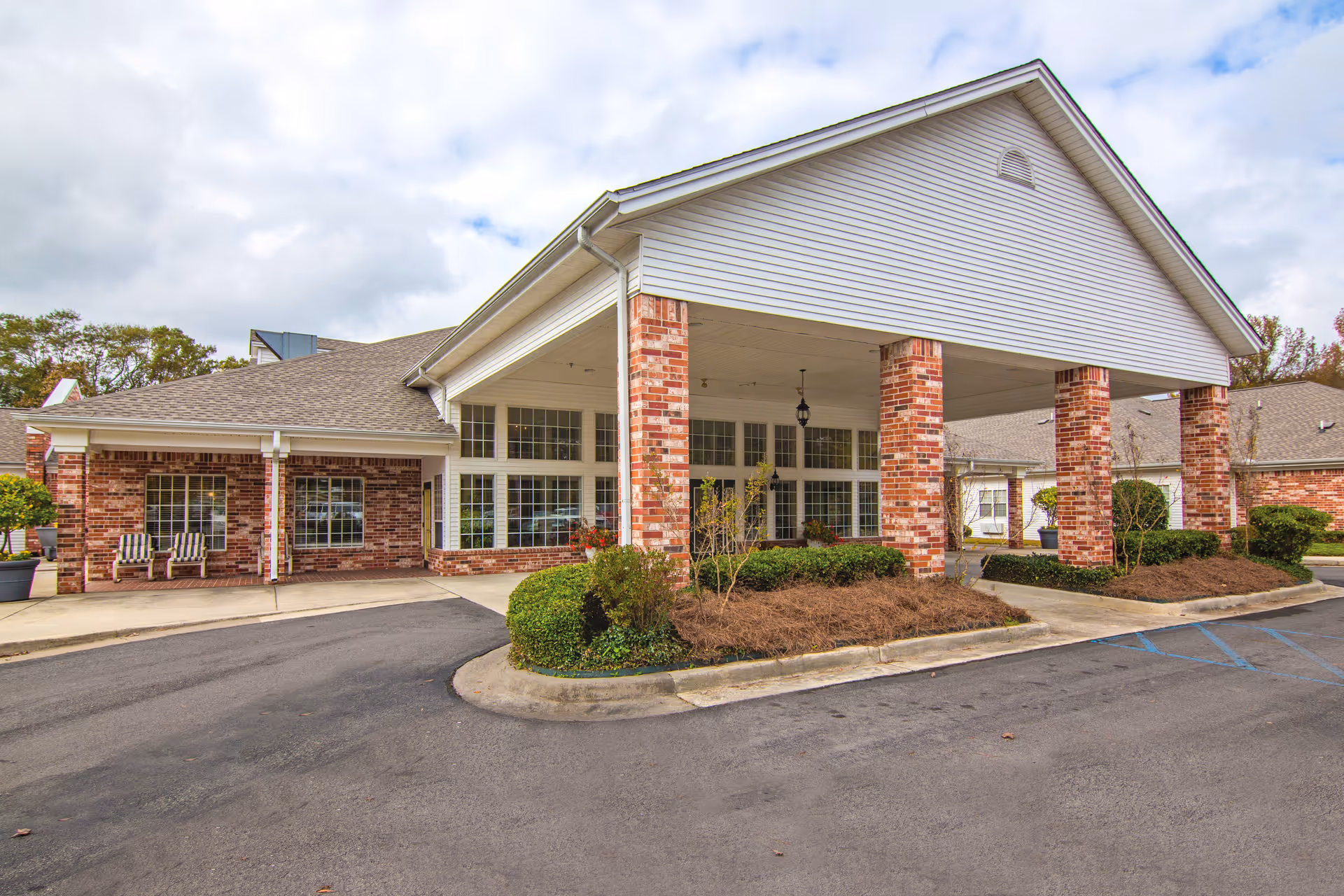 Exterior view of Bridgewood Gardens facility showing a brick building with large windows and a covered entrance supported by brick columns. There are landscaped bushes and a driveway in front with a cloudy sky overhead.