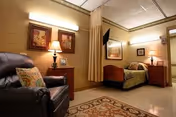 A cozy room with a single hospital-style bed, a bedside table with a lamp, a comfortable armchair with a decorative pillow, framed artwork on the walls, and a patterned rug on the floor. The room has warm lighting and a privacy curtain near the bed.