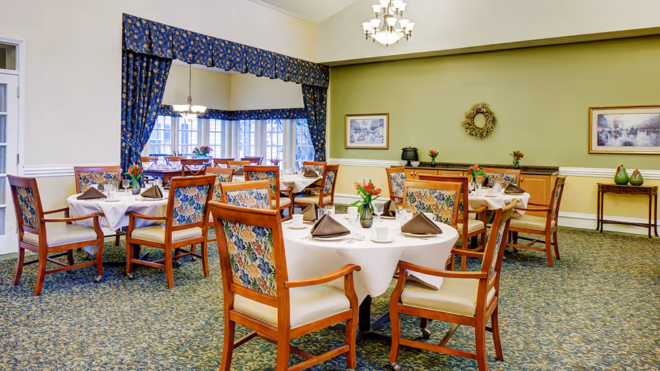 A dining room with several round tables covered with white tablecloths, each set with brown folded napkins, cups, and silverware. The chairs have wooden frames with floral upholstery. The room has green walls, carpeted floors, framed artwork, a wreath on the wall, and a window area with blue floral curtains.