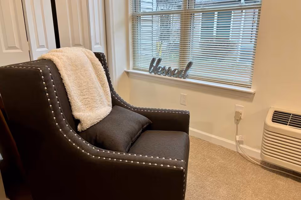 Dark gray studded armchair draped with a cream throw and cushion next to a window with blinds and a 'blessed' sign on the sill.