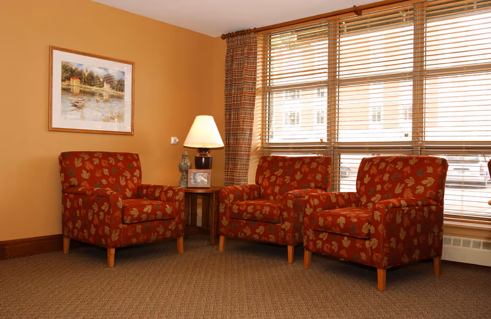 A cozy seating area with three red armchairs featuring a leaf pattern, arranged around a wooden side table with a lamp and decorative items. The room has beige walls, a large window with wooden blinds, and a framed painting on the wall.