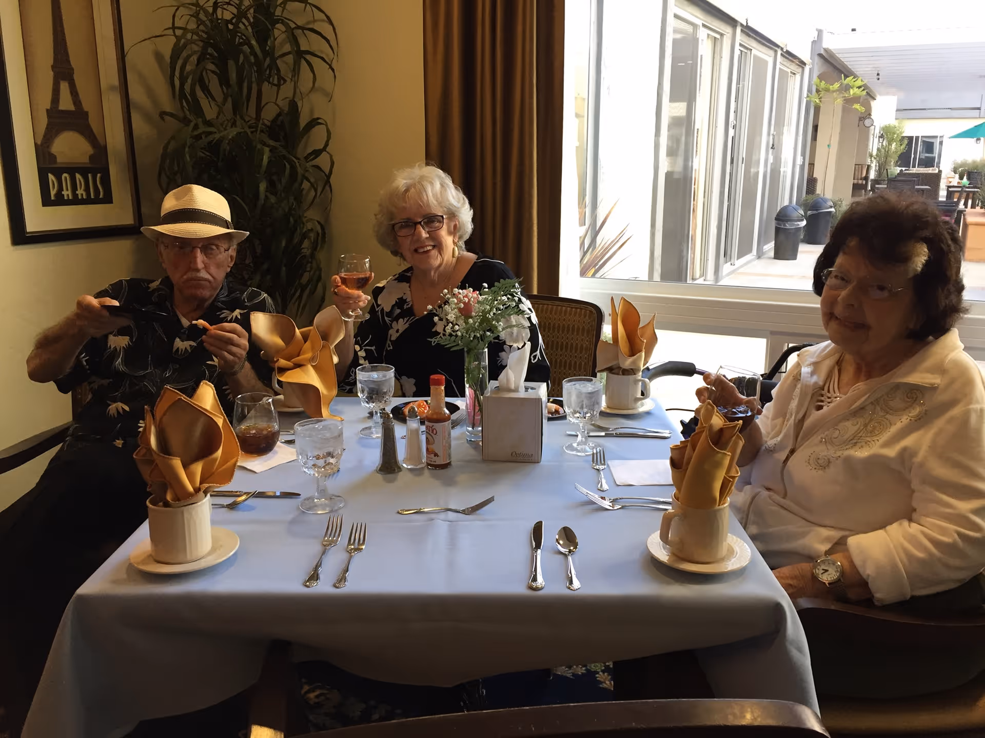 Three elderly people sitting at a dining table set with glasses, silverware, and napkins folded in cups. One woman in the center is holding up a glass and smiling, while the man on the left is eating and the woman on the right is seated with a slight smile. The room has a large window showing an outdoor patio area, a plant, and a framed picture of the Eiffel Tower with the word 'PARIS'.