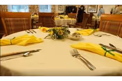 Communal dining room table set with yellow napkins, silverware, and a small floral centerpiece, with chairs and other tables in the background.