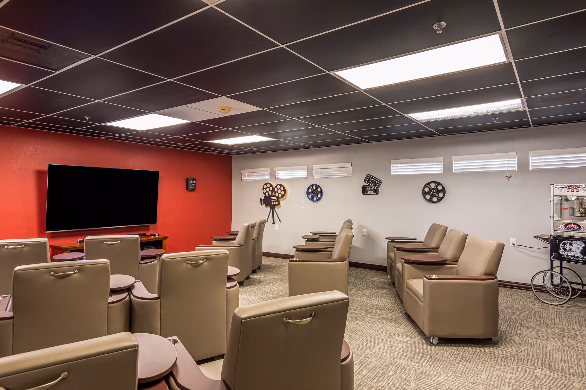 A small theater room with beige recliner chairs arranged in rows facing a large flat-screen TV mounted on a red accent wall. The opposite wall is decorated with vintage film reels and movie-themed decor. A popcorn machine is visible in the corner of the room.
