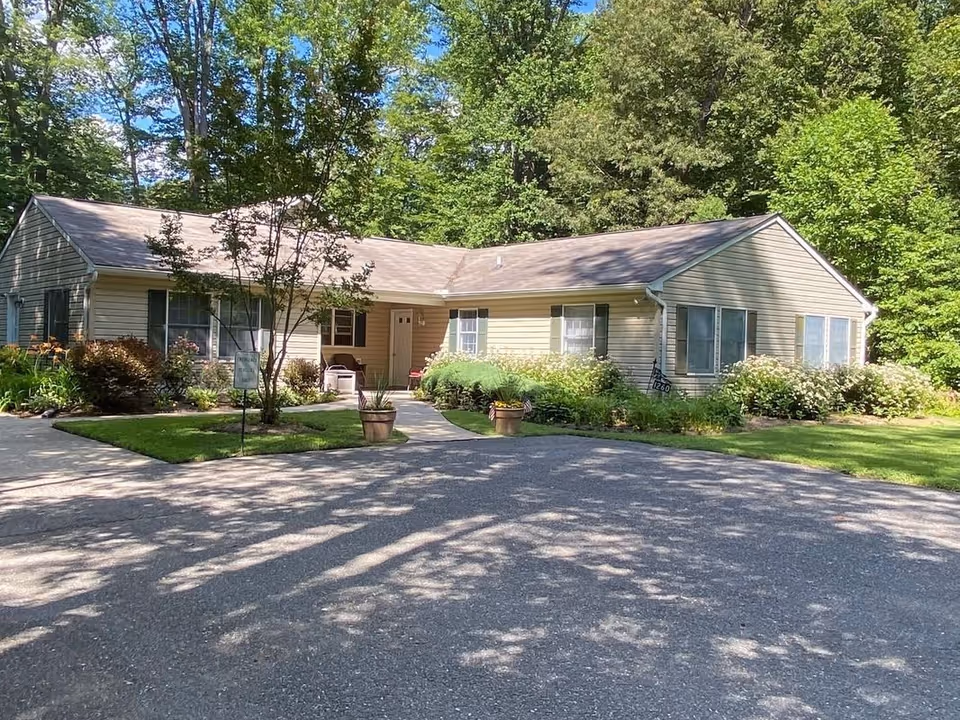 Single-story residential building with beige siding and green shutters surrounded by lush greenery and trees. There is a paved driveway and a small walkway leading to the front door, with potted plants and well-maintained bushes and flowers around the entrance.