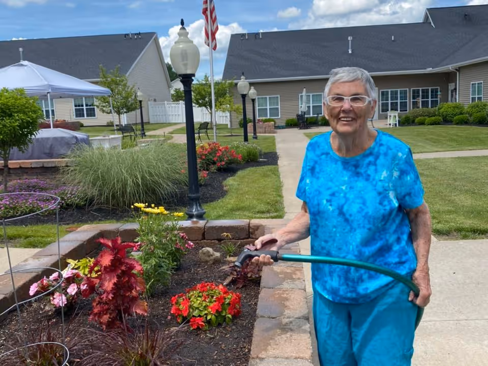 An elderly woman wearing a blue tie-dye shirt and blue pants is watering a flower bed with a green garden hose in an outdoor garden area of a senior living facility. Behind her are well-maintained lawns, flower beds, lamp posts, and single-story buildings under a partly cloudy sky.