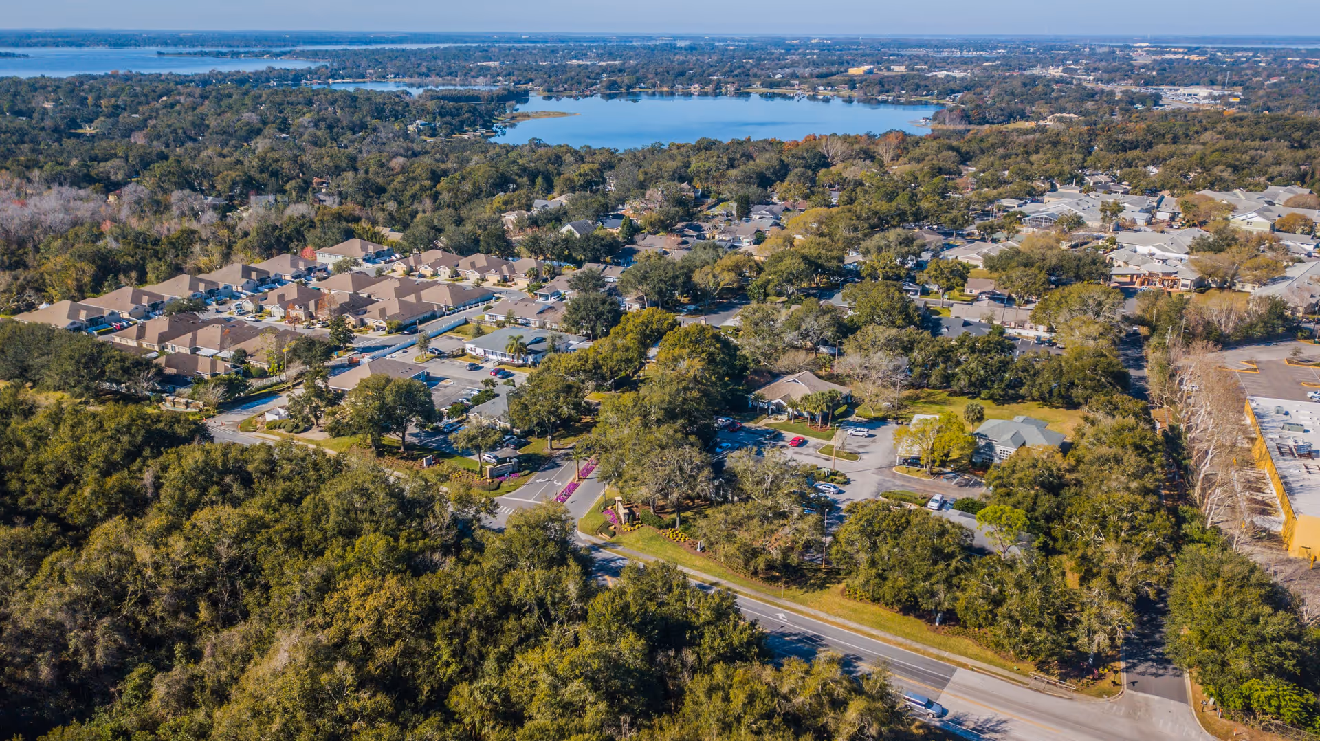 Aerial view of The Oaks at Waterman Village Retirement Community showing multiple residential buildings surrounded by trees and greenery, with a large body of water in the background and roads running through the area.
