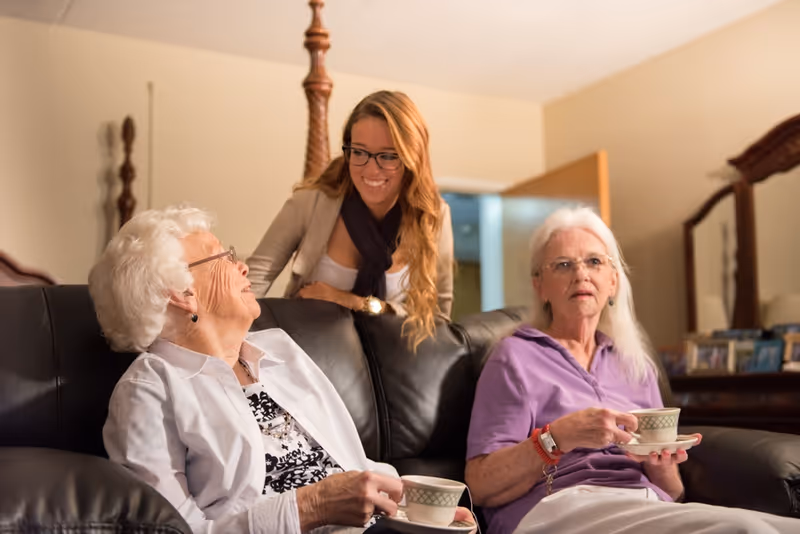 Two elderly women sitting on a black leather couch holding teacups, engaged in conversation with a younger woman standing behind the couch smiling at them in a cozy living room setting.