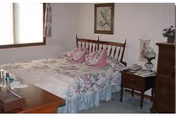 A bedroom with a wooden bed frame featuring a floral bedspread and matching pillows. There is a wooden nightstand with a lamp and a telephone, a dresser with decorative items, a window with blinds and floral curtains, and a framed picture hanging on the wall.