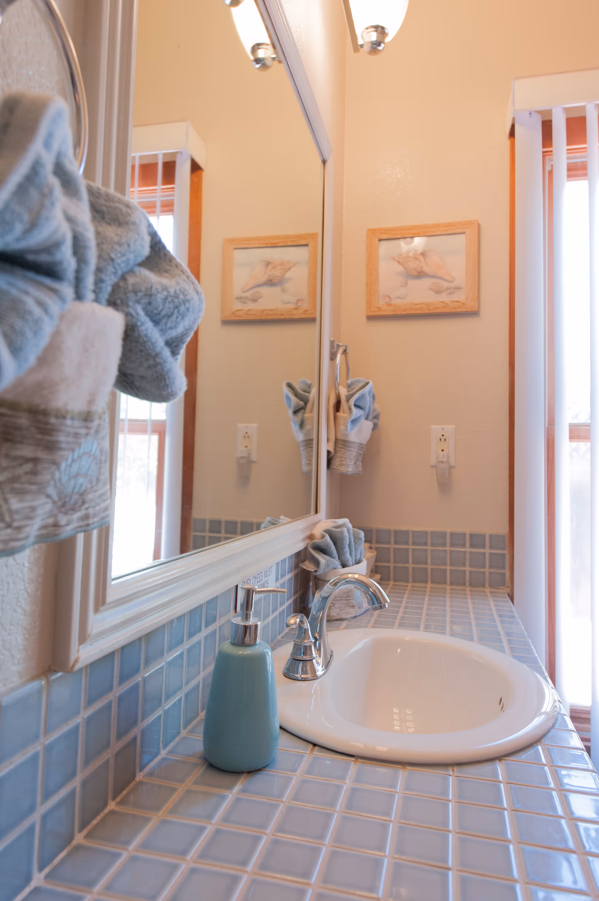 A bathroom sink area with blue tiled countertop and backsplash, a white oval sink with a silver faucet, a blue soap dispenser, and a mirror above the sink. A towel is hanging on the left side, and a framed picture is visible on the wall reflected in the mirror. There is a window with vertical blinds on the right side.
