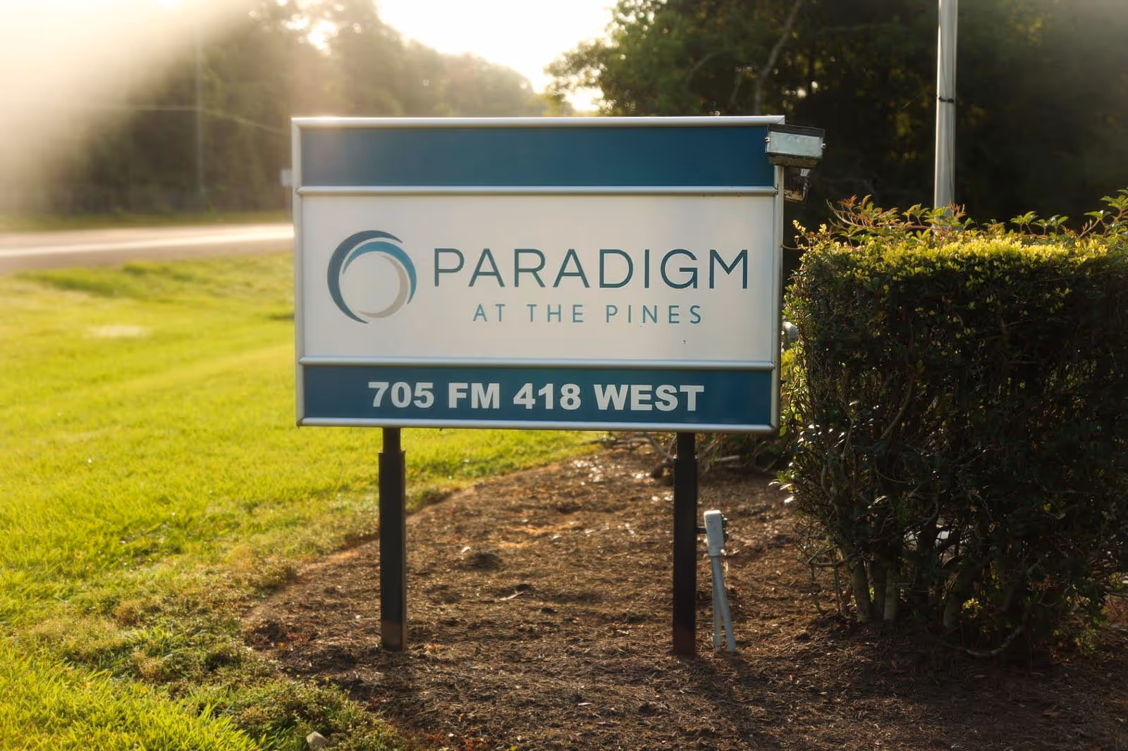 Outdoor sign for Paradigm at The Pines with the address 705 FM 418 West, surrounded by grass and bushes with sunlight in the background.