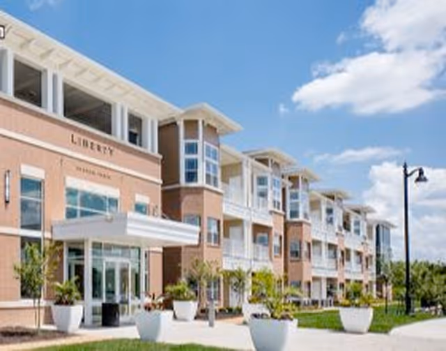 Exterior view of Liberty at Shoal Creek, a multi-story senior living facility with large windows, balconies, and a covered entrance. The building is surrounded by landscaped greenery and large white planters under a partly cloudy blue sky.