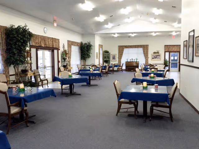 A spacious dining room with multiple tables covered in blue tablecloths, each surrounded by chairs. The room has large windows with curtains, potted plants, framed pictures on the walls, and a piano at the far end. The ceiling has recessed lighting, and the floor is carpeted.