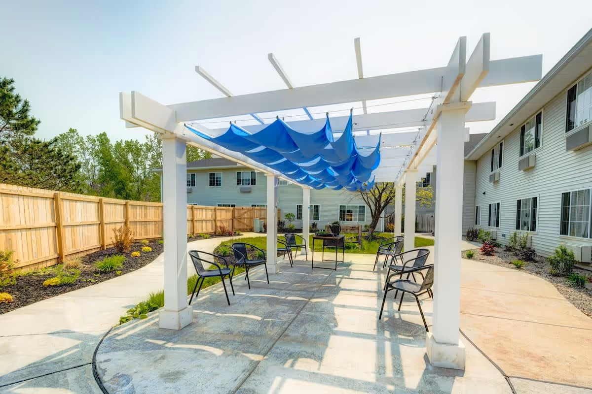 Outdoor patio area at Evergreen Place featuring a white pergola with blue fabric shades overhead, surrounded by black metal chairs arranged in a circle on a concrete surface. The area is enclosed by a wooden fence with landscaping along the edges and residential buildings in the background.