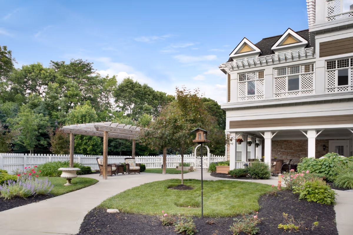 Outdoor garden area at Sunrise of Dublin featuring a paved walkway, a wooden pergola with seating, manicured lawns, flower beds, and a multi-story building with a covered porch in the background under a partly cloudy sky.