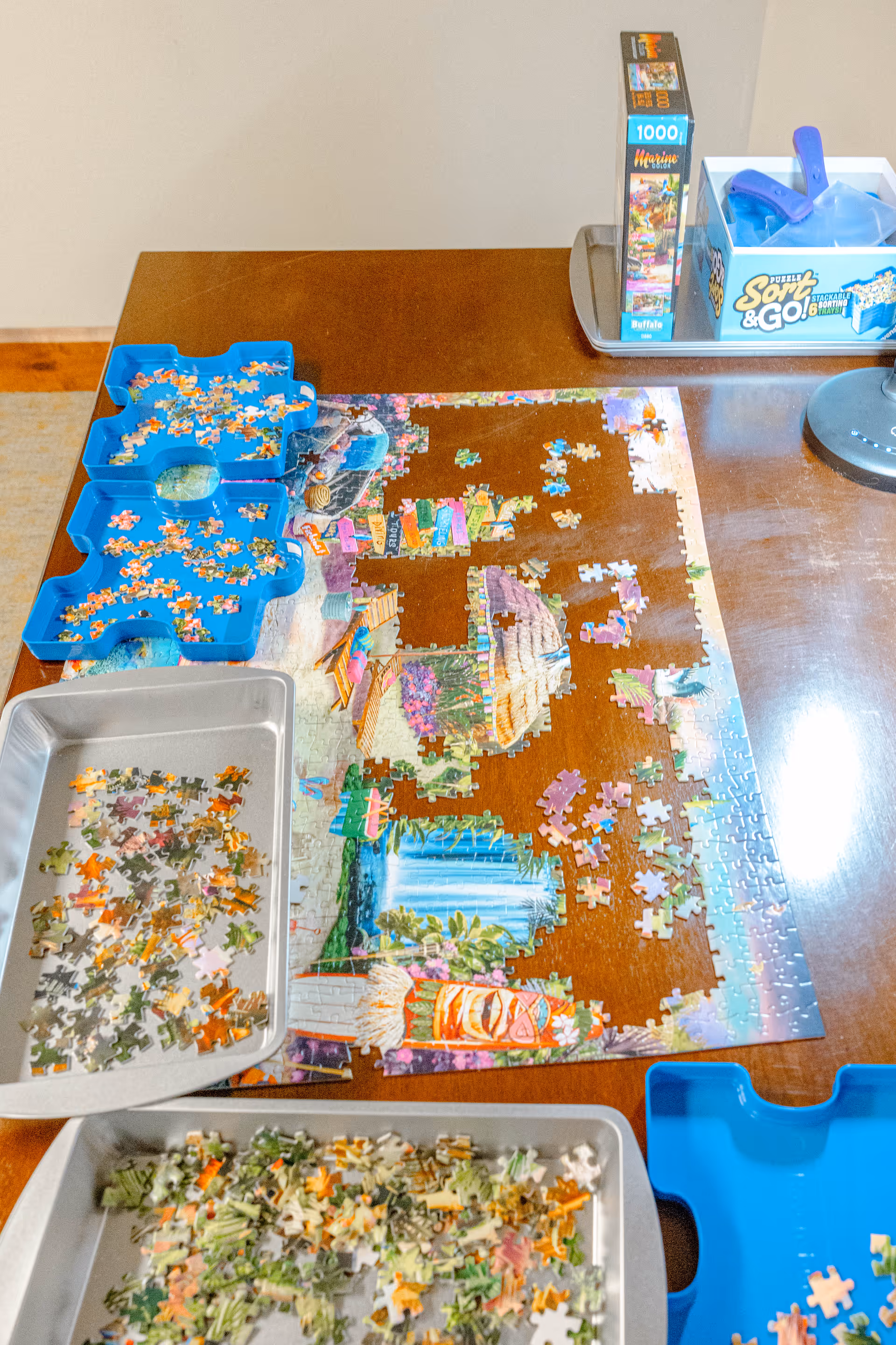A partially completed jigsaw puzzle on a wooden table with several puzzle pieces sorted into blue and silver trays. A box labeled 'Sort & Go!' and a 1000-piece puzzle box are visible on the table.