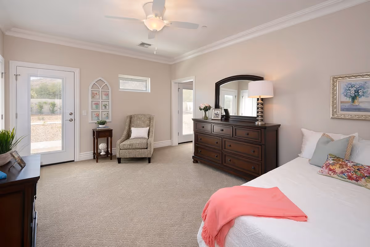 A bright and spacious bedroom with beige walls and carpet. The room features a large bed with white bedding, colorful pillows, and a coral throw blanket. There is a dark wooden dresser with a large mirror and a table lamp on top. A comfortable armchair with a white pillow sits next to a small wooden side table with a plant. Two doors with glass panels lead outside, and a small window is above the armchair. The room is decorated with framed artwork and has a ceiling fan with a light.