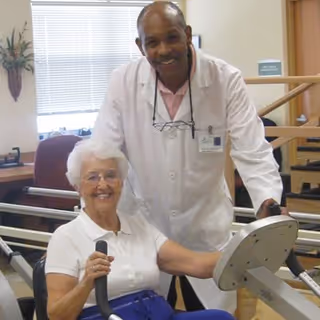 An elderly woman using a seated exercise machine in a health care facility, assisted by a male healthcare professional wearing a white coat and a name badge. They are both smiling and appear to be in a rehabilitation or fitness area with exercise equipment and handrails visible in the background.