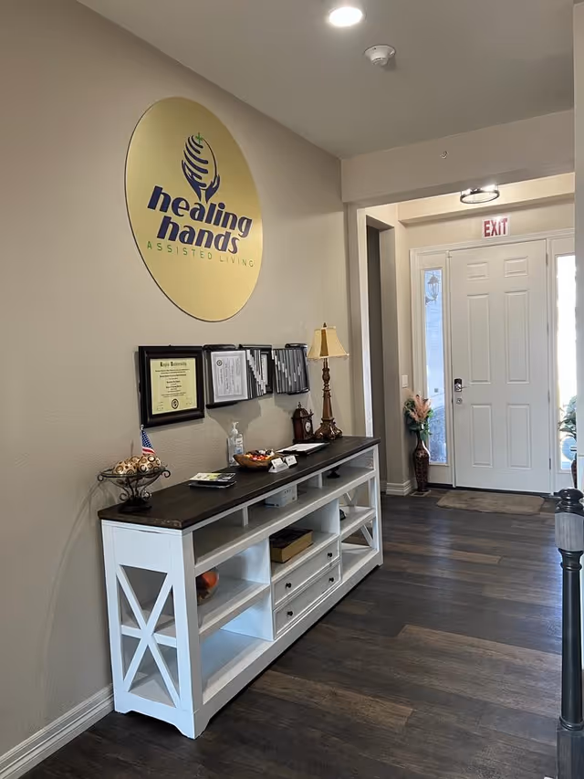 Interior view of a hallway in Healing Hands Assisted Living featuring a white console table with a dark wooden top against a beige wall. On the wall above the table is a large round sign with the facility's logo and name. The table holds decorative items, a lamp, hand sanitizer, and some documents. At the end of the hallway is a white door with an exit sign above it and a vase with flowers beside it.