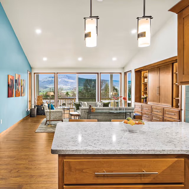 Bright and spacious living room area with large windows offering a scenic view of mountains and greenery. The room features wooden flooring, a teal accent wall with colorful artwork, comfortable seating including a sofa and armchairs, and a wooden built-in cabinet. In the foreground, there is a kitchen island with a marble countertop and a bowl of fruit, illuminated by two hanging pendant lights.