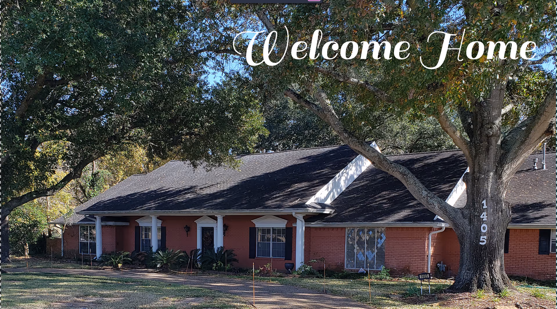 A single-story brick building with a dark roof surrounded by large trees and greenery. The building has white columns at the entrance and multiple windows. The words 'Welcome Home' are overlaid at the top of the image, and the number 1405 is visible on a tree in the foreground.