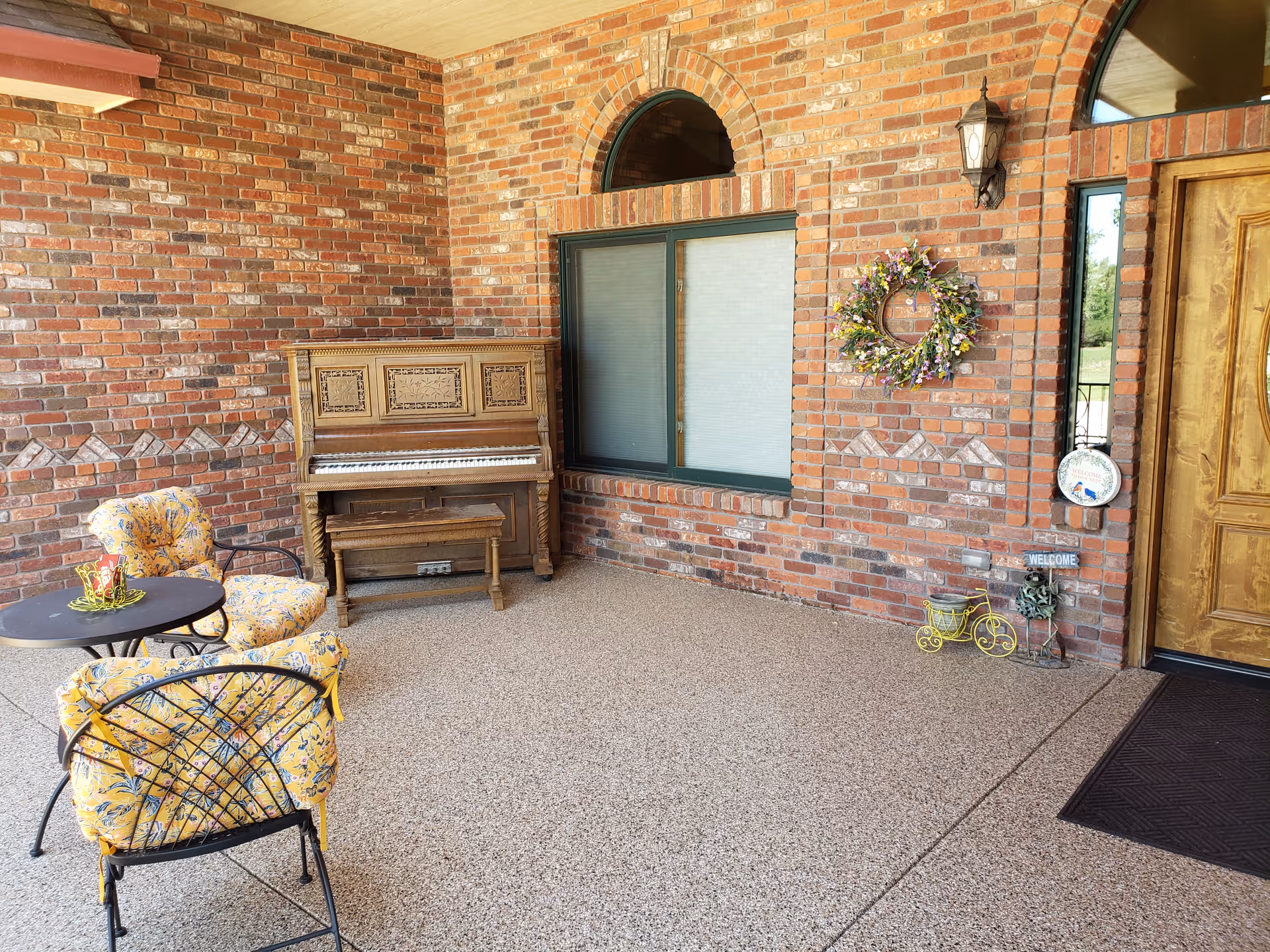 A covered outdoor patio area with brick walls, featuring a wooden upright piano, two cushioned chairs with yellow floral upholstery, a small round black table, a decorative wreath on the wall, a wooden door, and various small decorative items including a welcome sign and a miniature yellow bicycle.