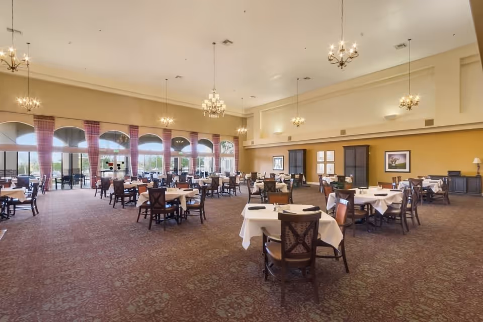 Spacious dining room with multiple tables covered in white tablecloths and set with napkins and utensils. The room features large arched windows with red plaid curtains, chandeliers hanging from a high ceiling, and carpeted floors. There are cabinets and framed artwork on the yellow walls.