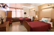 A bright shared bedroom with two single beds, bedside tables, chairs, a dresser, and a wall-mounted TV.