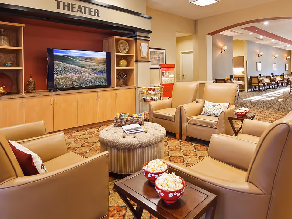 A cozy theater room with four tan leather armchairs arranged around a small table holding two bowls of popcorn. A large flat-screen TV is mounted on a wooden cabinet with shelves displaying decorative items. The word 'THEATER' is displayed above the TV. In the background, there is a popcorn machine and a dining area with tables and chairs.