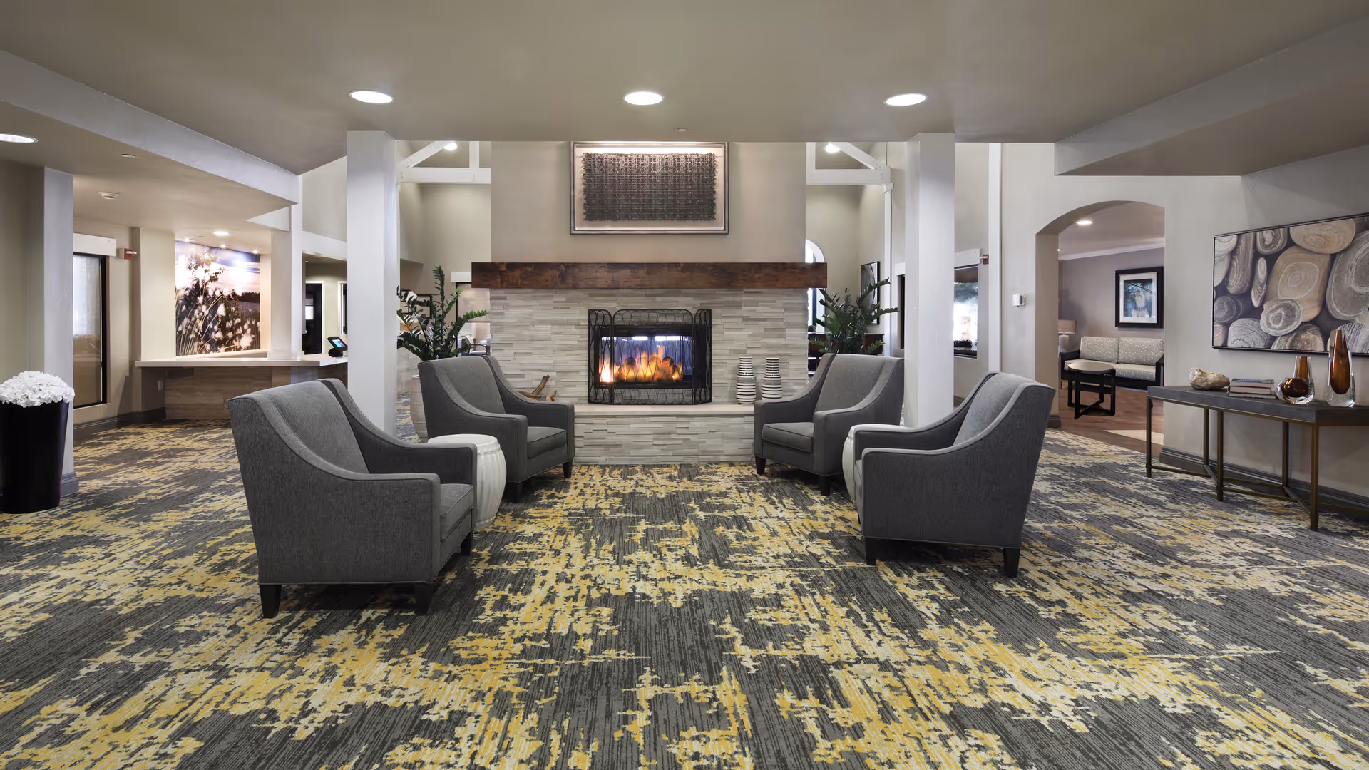 A spacious and modern living room area in a senior living facility featuring a central fireplace with a wooden mantel, surrounded by four gray upholstered armchairs arranged in a square. The room has a patterned carpet with shades of gray and yellow, white pillars, and neutral-colored walls. Decorative plants and artwork are visible in the background, along with additional seating areas.