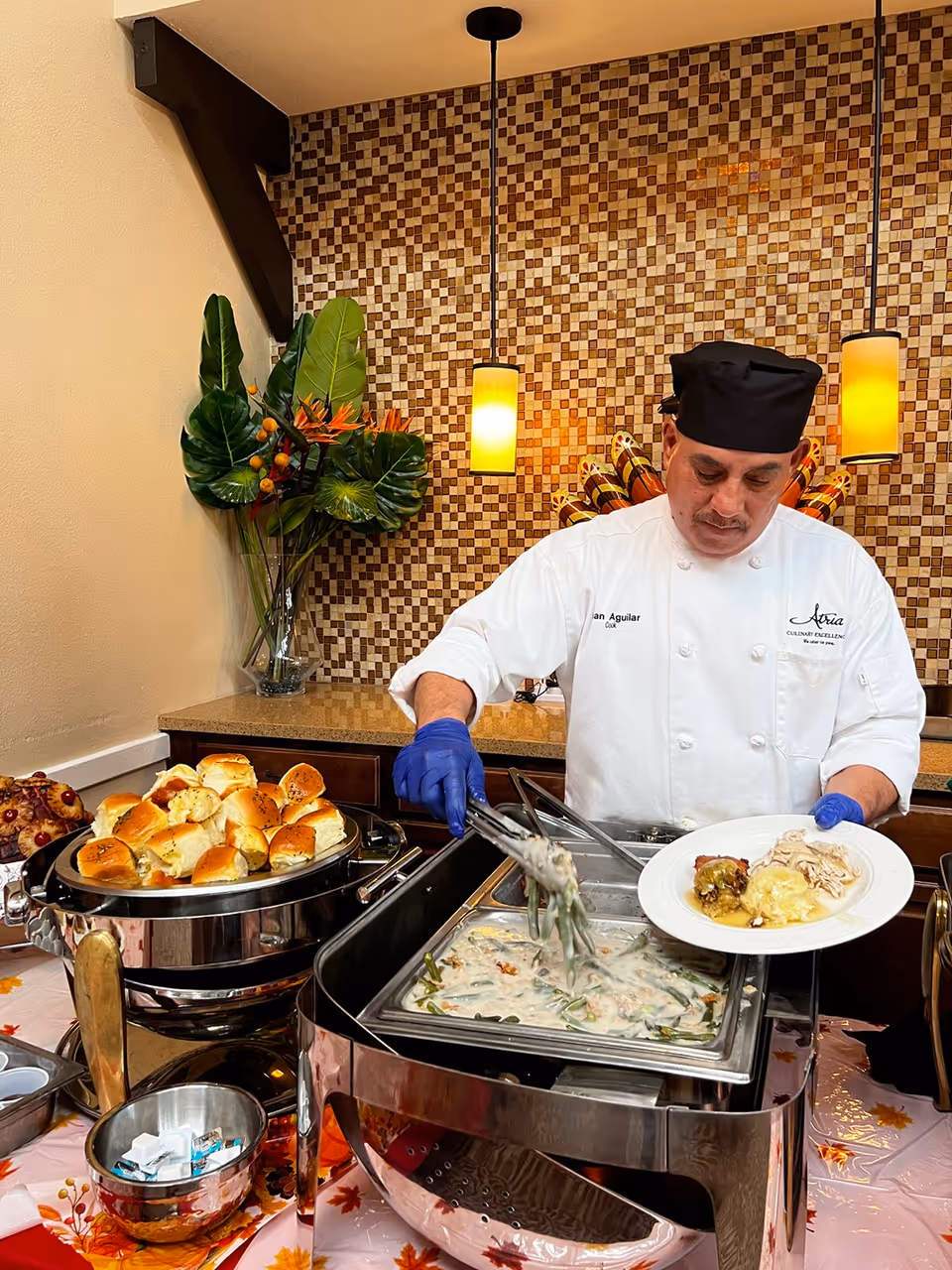 A chef wearing a white uniform and black hat serves food onto a plate from a buffet station. The buffet includes a dish with green beans in a creamy sauce and a tray of dinner rolls. The background features a mosaic tile wall and two hanging pendant lights, with a vase of tropical leaves and flowers on the counter.