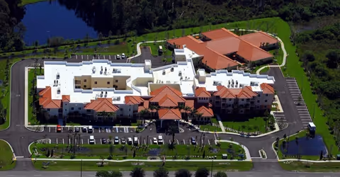 Aerial view of Discovery Village At The Forum senior living facility showing a large building complex with white and terracotta-colored roofs, surrounded by parking lots, green lawns, and trees, with a pond visible in the background.