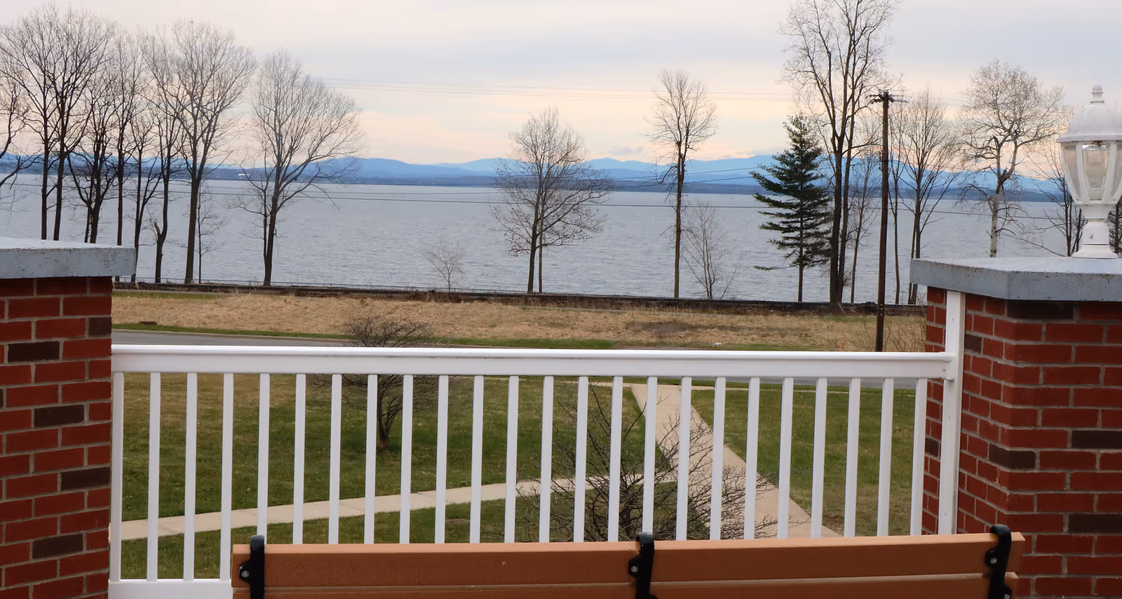 View from a brick balcony with a white railing and bench overlooking grass, bare trees and a lake with distant hills.