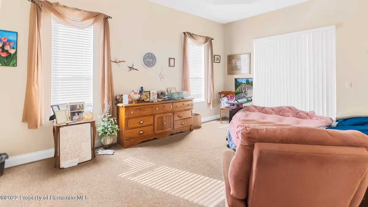 A cozy living room area in an assisted living facility with beige walls and carpet. The room features two windows with beige curtains, a wooden dresser with various framed photos and decorative items on top, a small table with more framed photos and a whiteboard, a pink armchair, and a bed covered with a pink blanket. There is also a TV on a stand near the corner and a chair beside it. Vertical blinds cover a large window or sliding door on the right side.