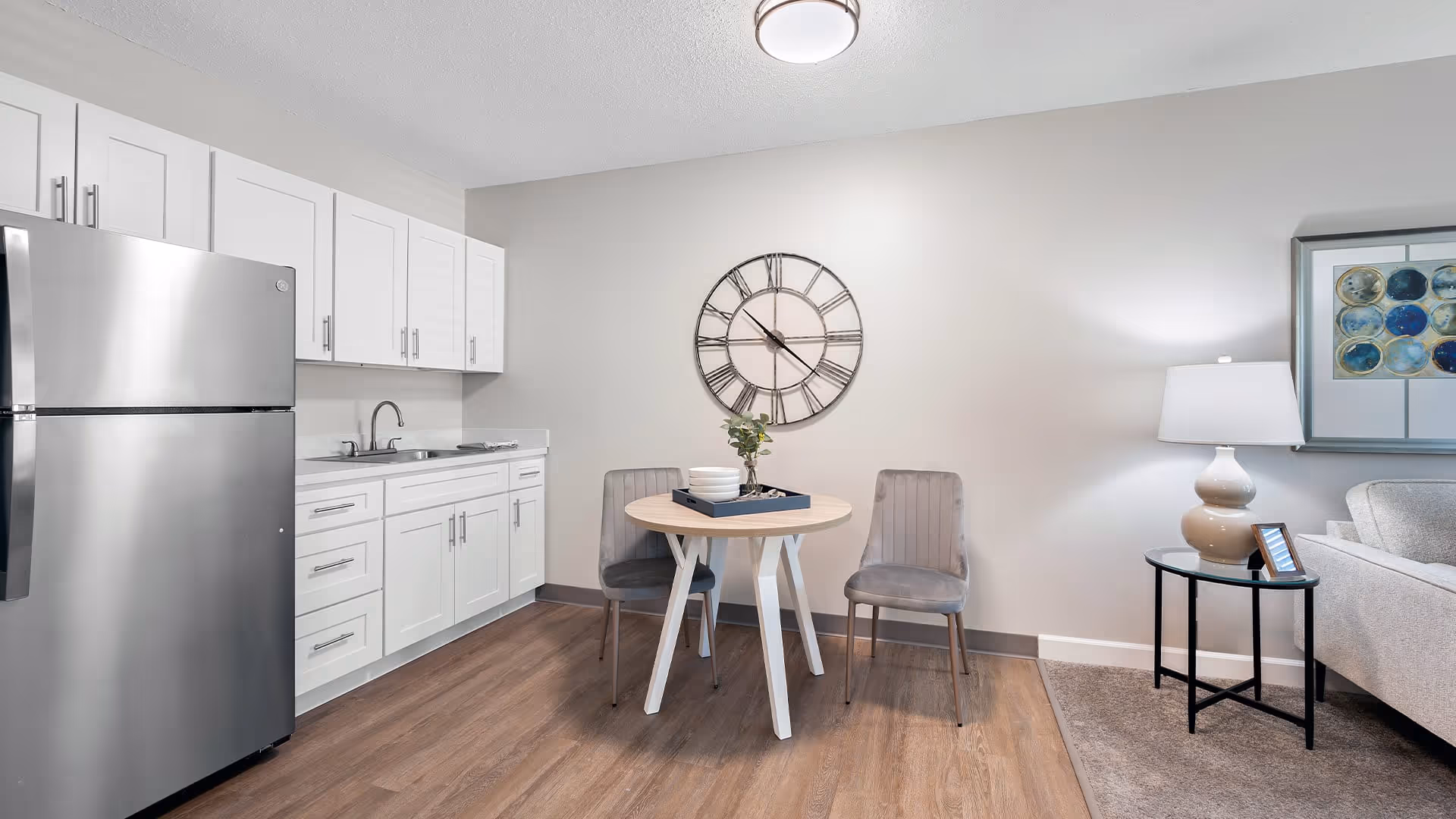 A modern kitchen and dining area with white cabinets, a stainless steel refrigerator, a small round wooden table with two gray chairs, a large wall clock, and a side table with a lamp and framed picture next to a light-colored sofa.