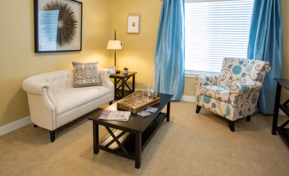 A cozy living room with a beige tufted loveseat with a decorative pillow, a patterned armchair with blue and beige floral designs, a black coffee table with a wooden tray holding glassware and magazines, a side table with a lamp, small plant, and picture frame, yellow walls, blue curtains, and a window with blinds.