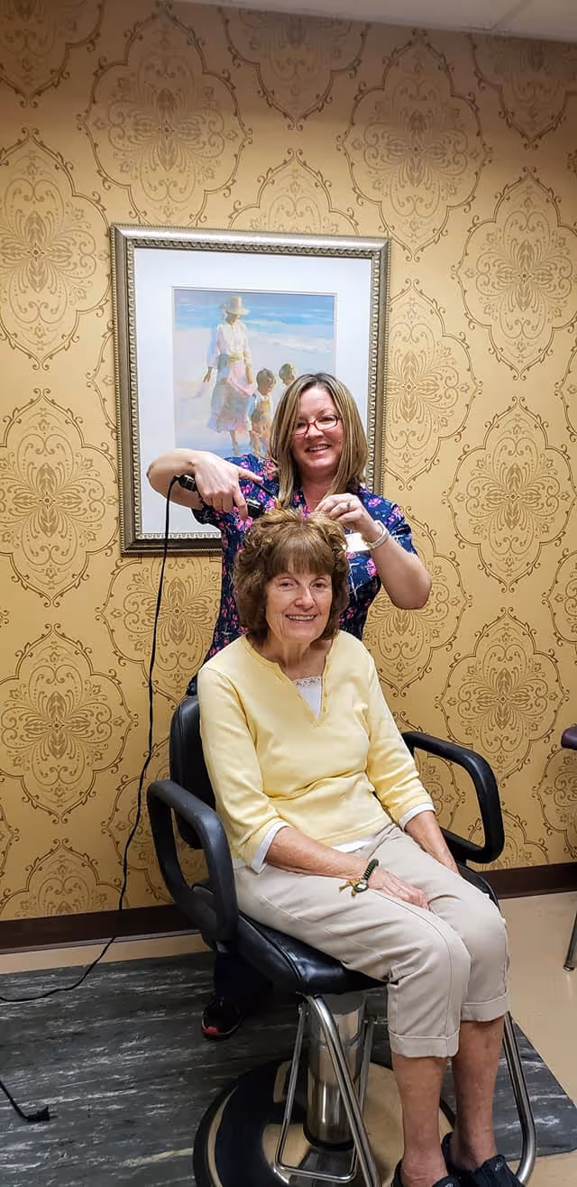A woman with short brown hair wearing a yellow top and light-colored pants is seated in a black salon chair. Another woman standing behind her, wearing glasses and a floral top, is styling her hair with a curling iron. The room has ornate gold patterned wallpaper and a framed painting of a woman and children on the wall behind them.