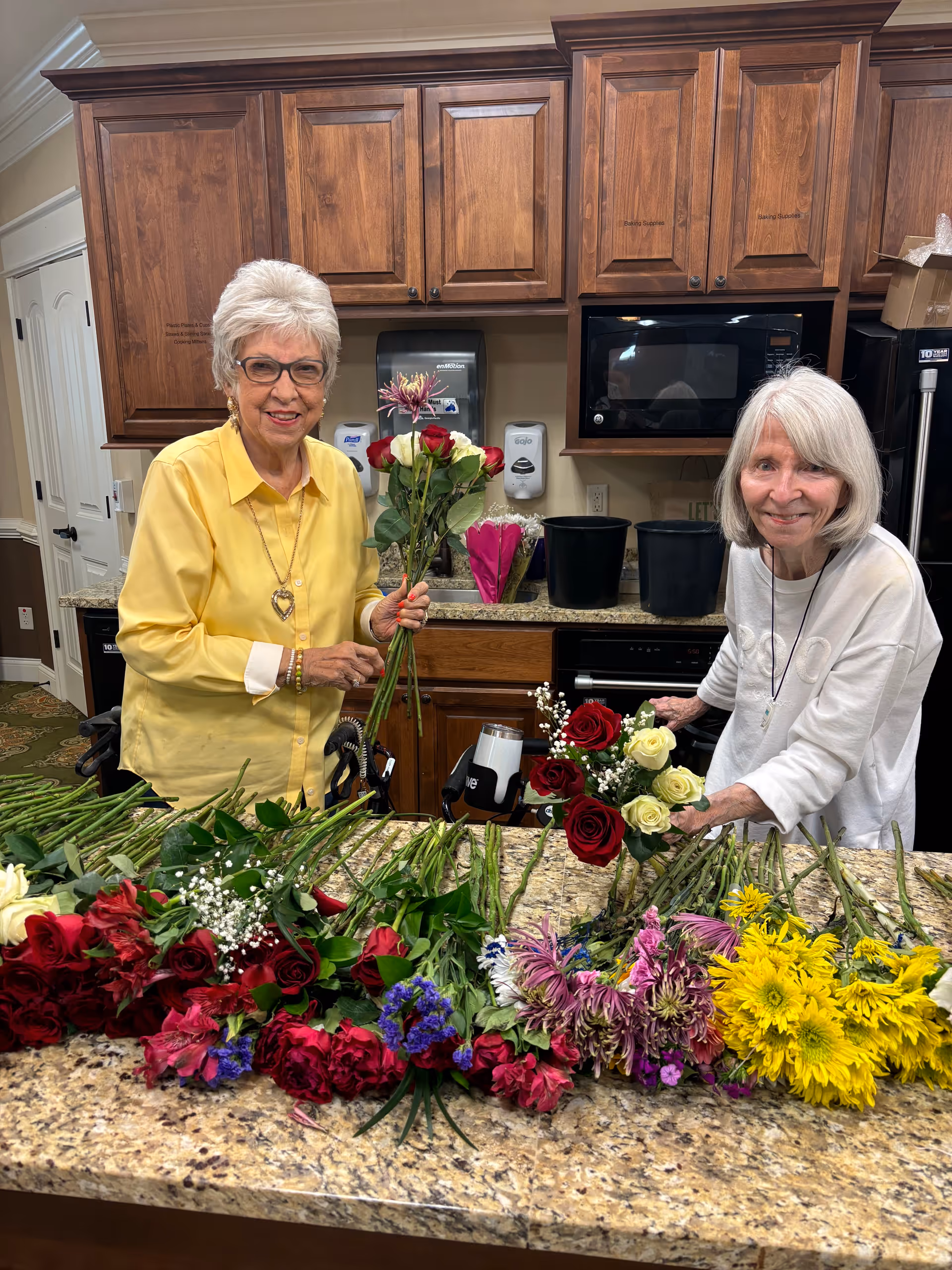 Two elderly women arranging various colorful flowers on a kitchen counter in a room with wooden cabinets and a microwave. One woman is wearing a yellow shirt and glasses, holding a bouquet of red and white flowers. The other woman is wearing a white shirt and smiling while holding a bouquet of red and white flowers.
