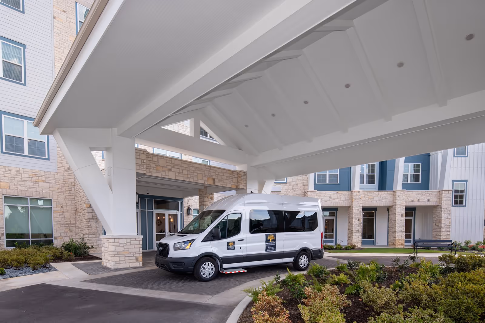 Exterior view of The Summit of Germantown senior living facility entrance with a covered drop-off area and a white shuttle van parked underneath. The building features stone and siding exterior with multiple windows and landscaped greenery around the driveway.