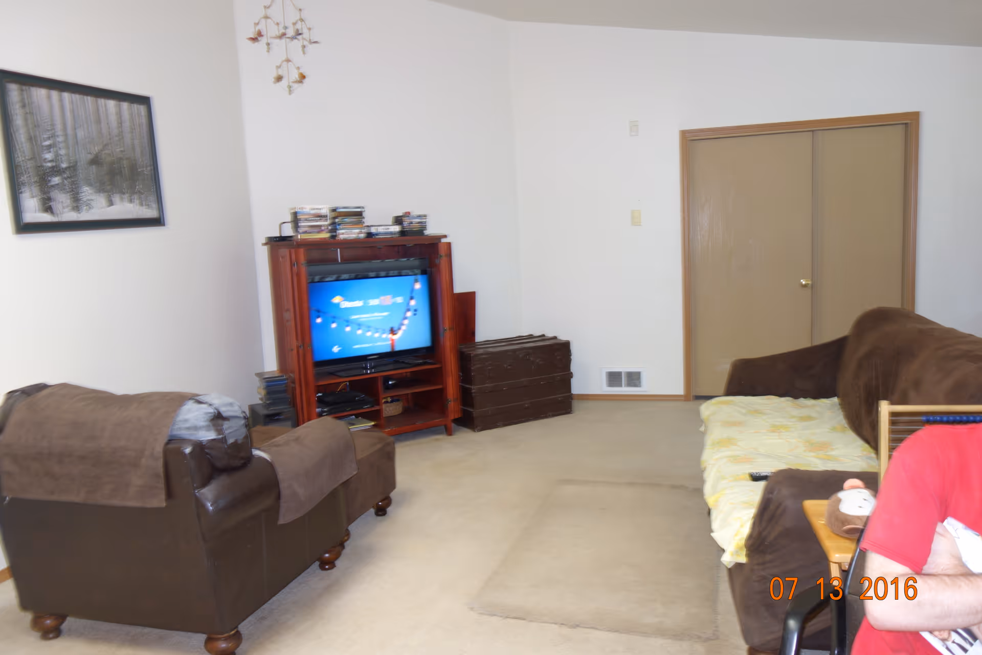 A living room with a brown armchair and a brown sofa covered with a yellow blanket. There is a wooden TV stand with a flat-screen TV and stacks of DVDs or CDs on top. A framed picture hangs on the white wall, and there is a closed double door in the background. A person wearing a red shirt is partially visible sitting on the right side.