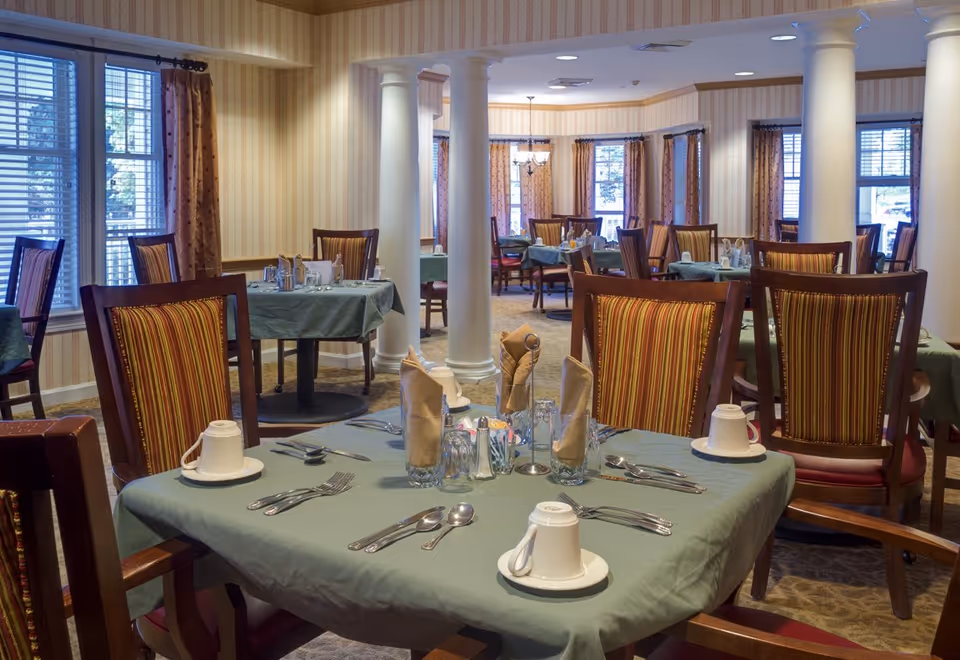 Interior view of a dining room in a senior living facility with tables covered in green tablecloths, set with cups, glasses, silverware, and napkins. The room features striped upholstered chairs, large windows with curtains, and white columns.